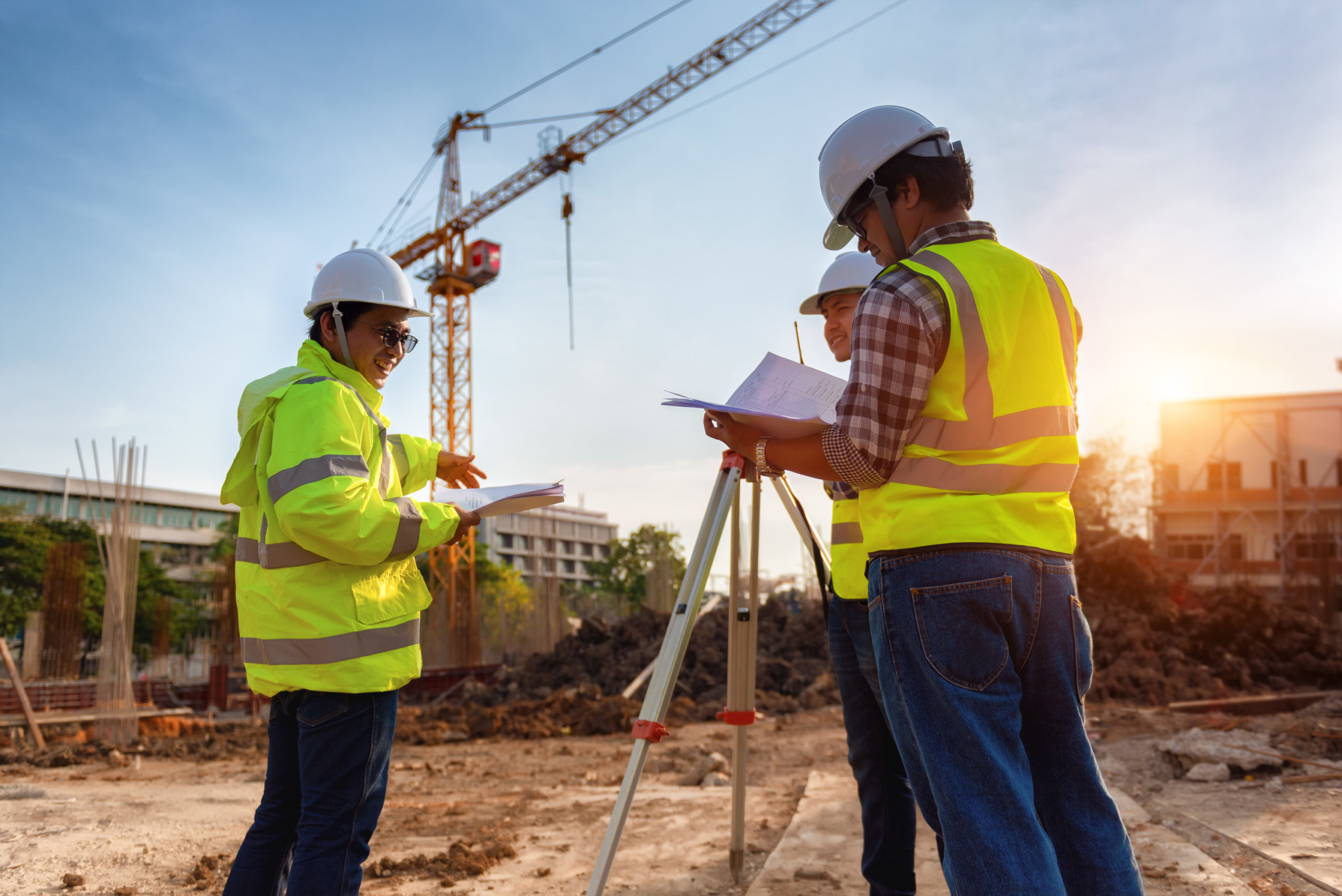 cropped-AdobeStock_249447070_50-scaled-1.jpeg Surveyors in hard hats and safety vests review plans beside a tripod at a construction site